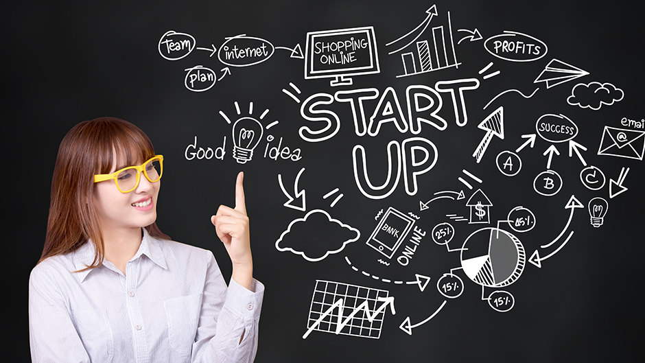Woman in front of a chalkboard that say Start Up and has business-related image drawn on it